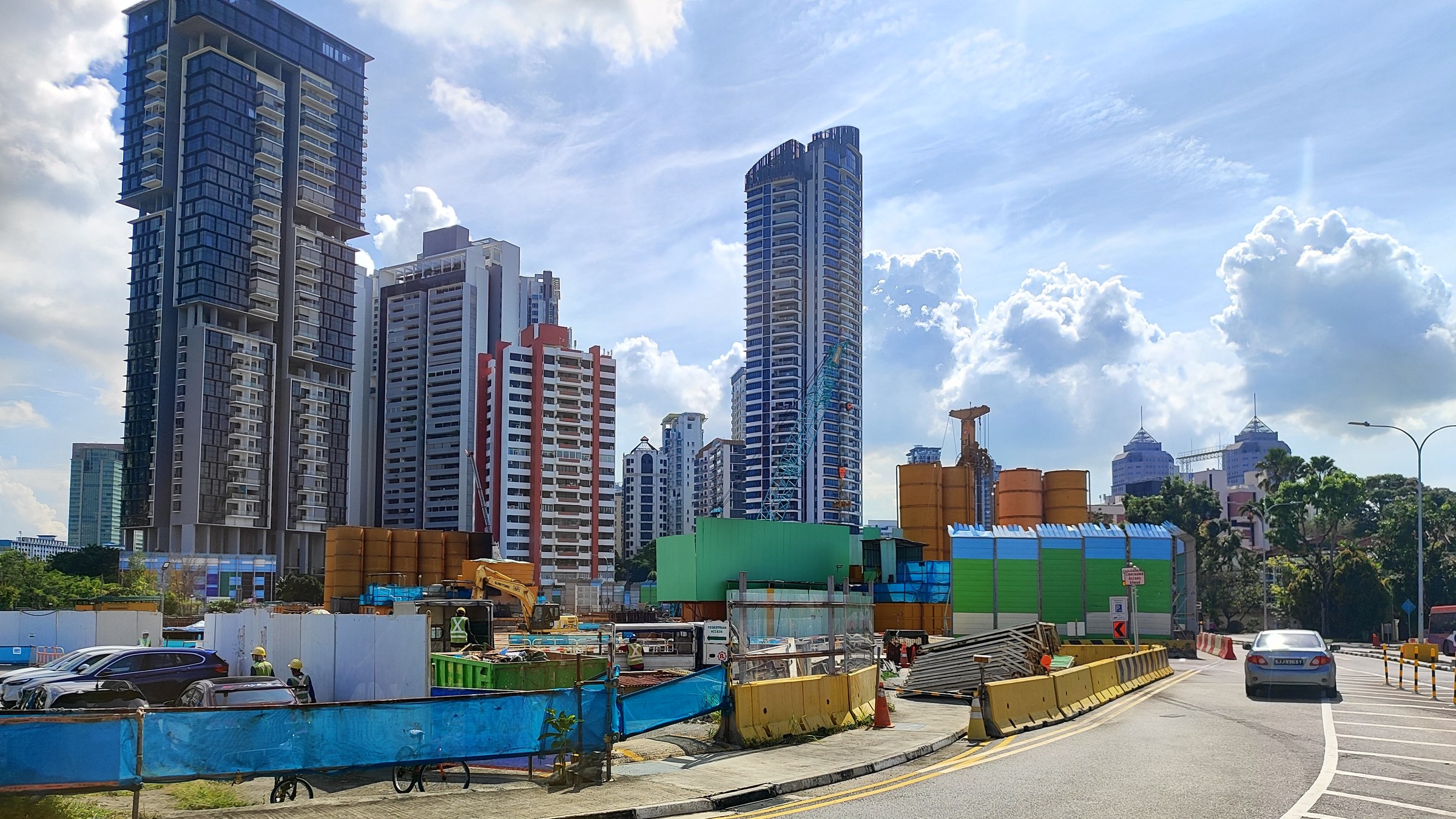 Construction hoardings and machinery for the North-South Corridor along Thomson Road, with high-rise condominium towers and blue sky behind.