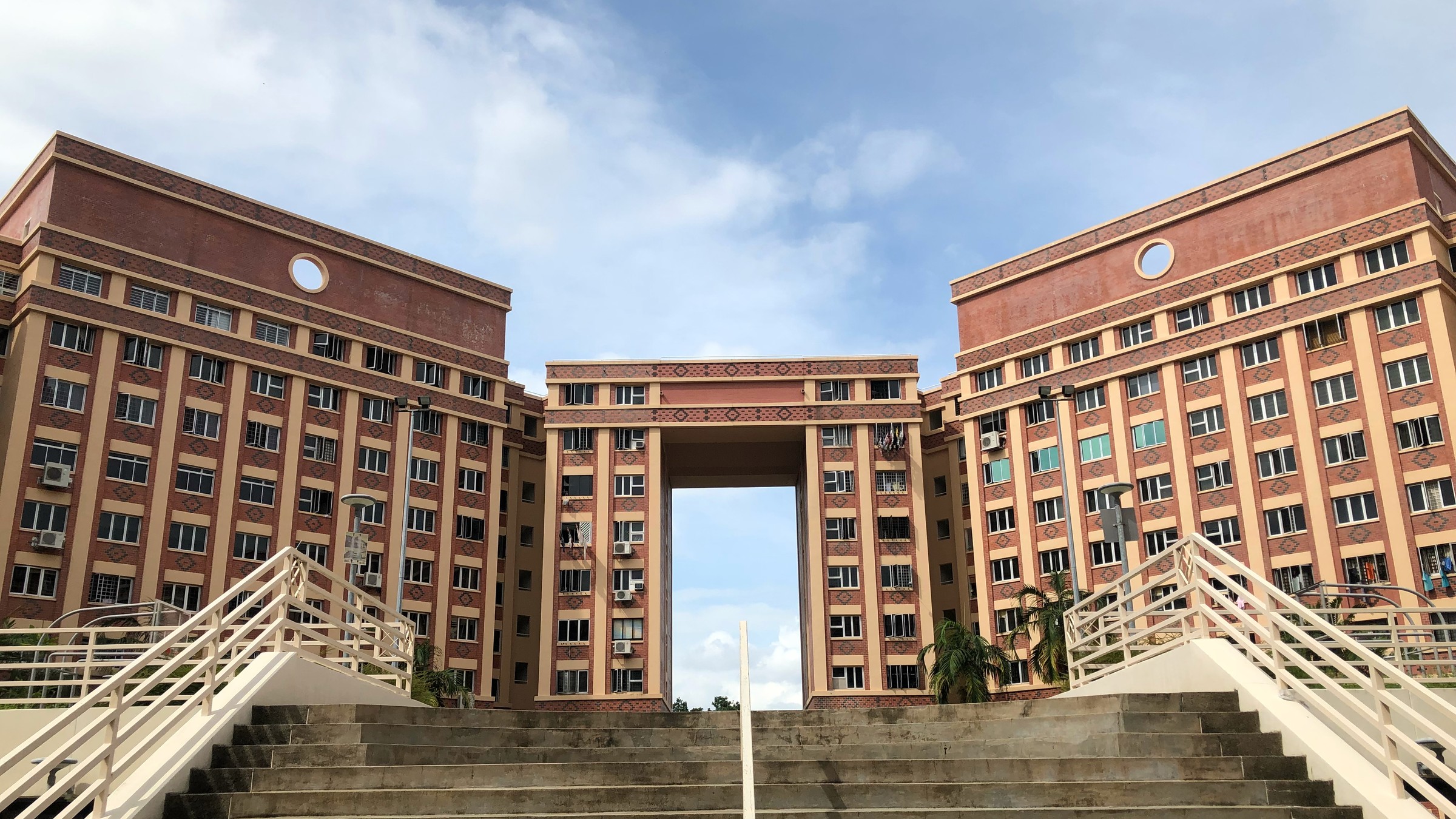 Red-brick HDB blocks in Tampines framing a central archway, seen from the top of a wide staircase under a blue sky