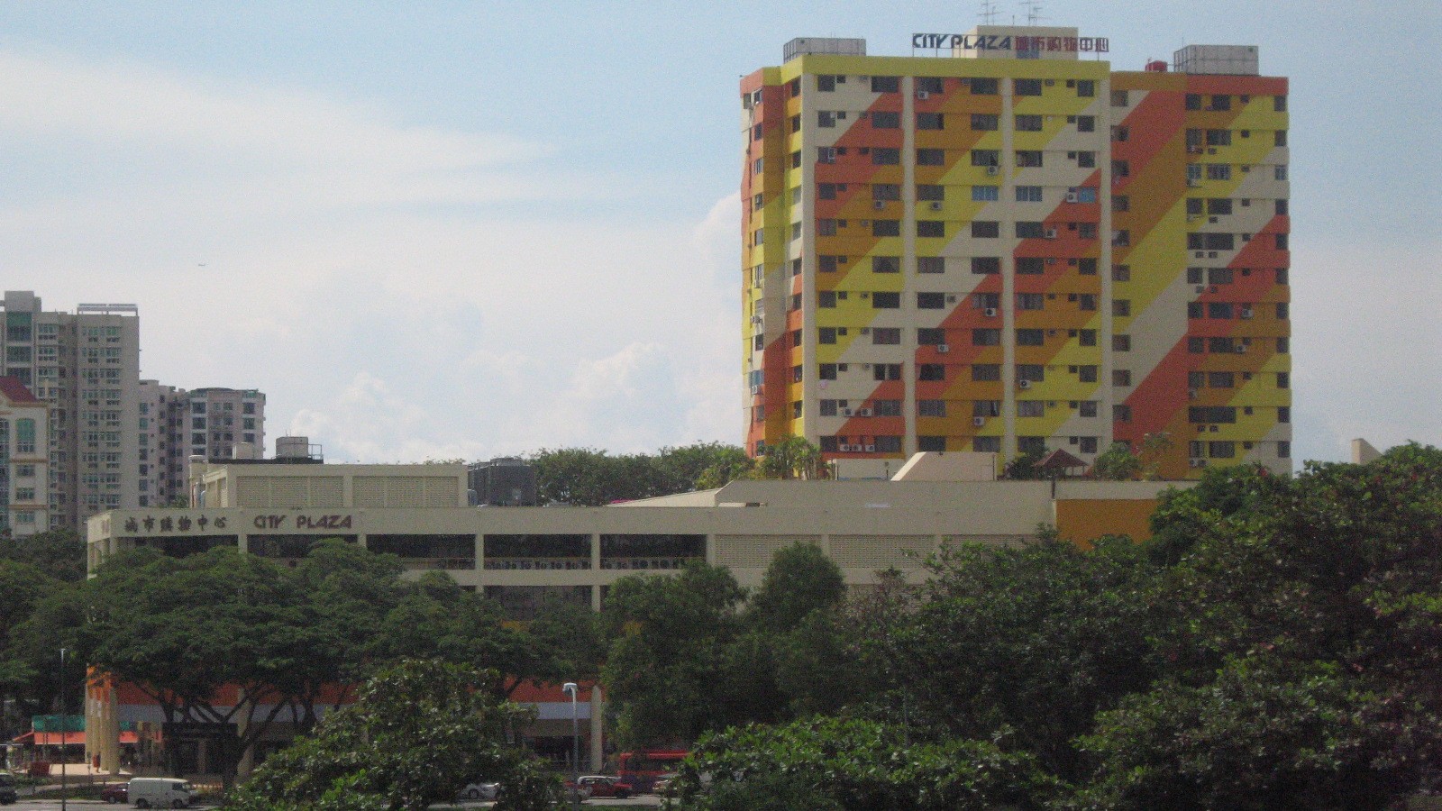 City Plaza, an 18-storey tower in orange, yellow and cream stripes above a long retail podium, seen across trees from the Geylang Road side, with newer condominiums in the background.