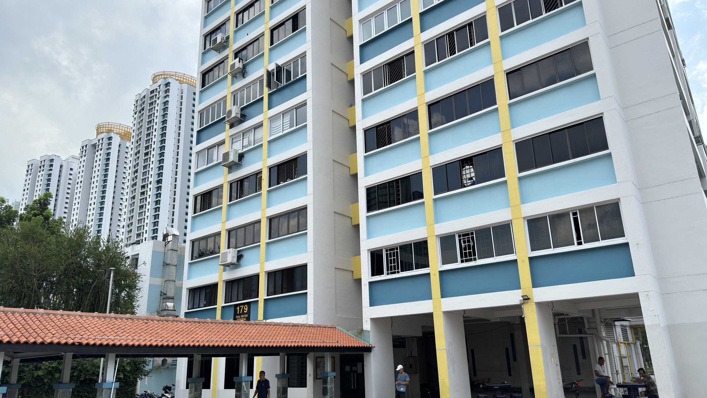 An HDB block in Toa Payoh with a sheltered walkway and residents at the void deck, with taller blocks behind.