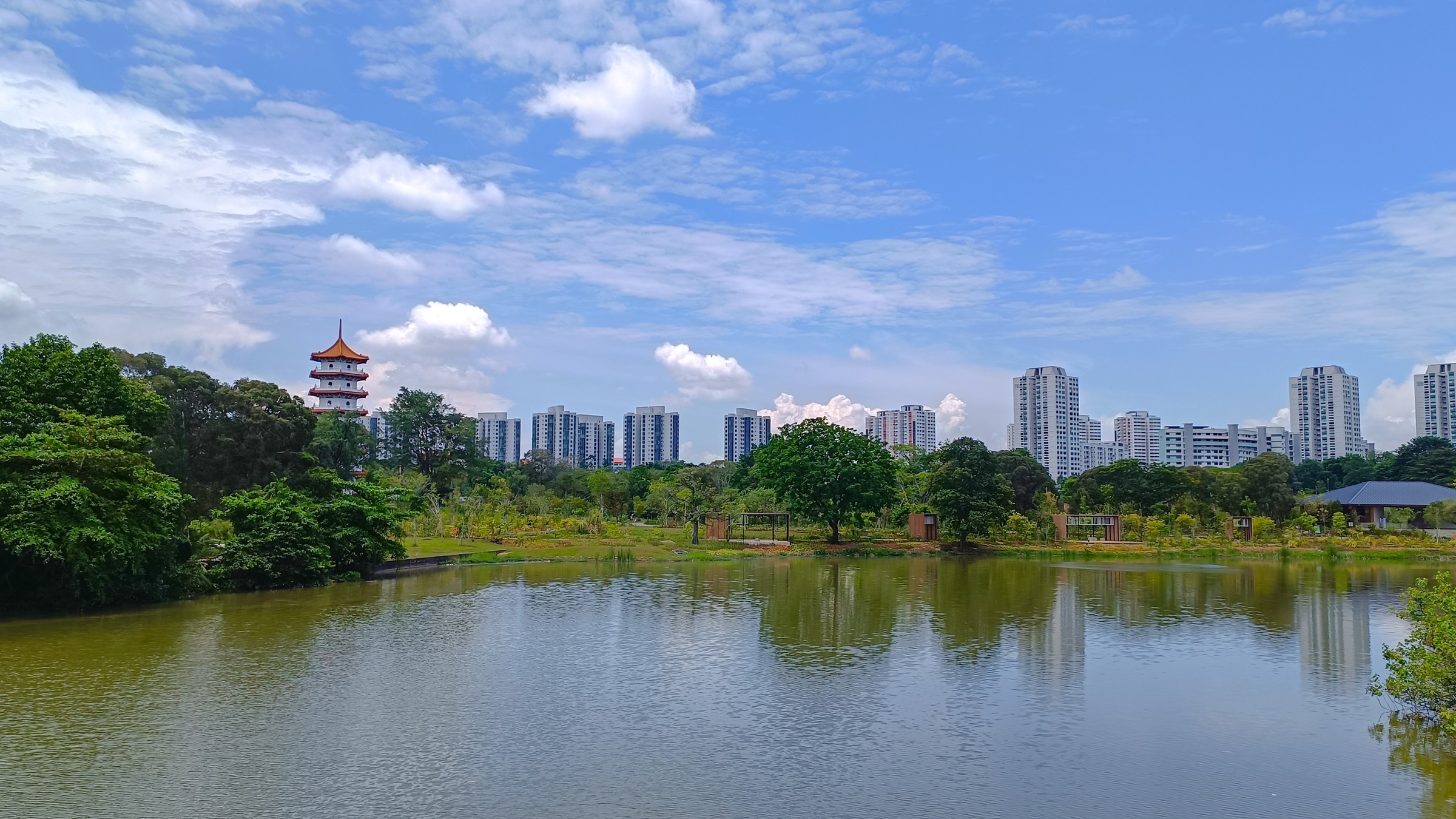 Jurong Lake seen from the Moon Bridge in Jurong Lake Gardens, with the Chinese Garden pagoda on the left and residential towers lining the horizon under a bright blue sky.