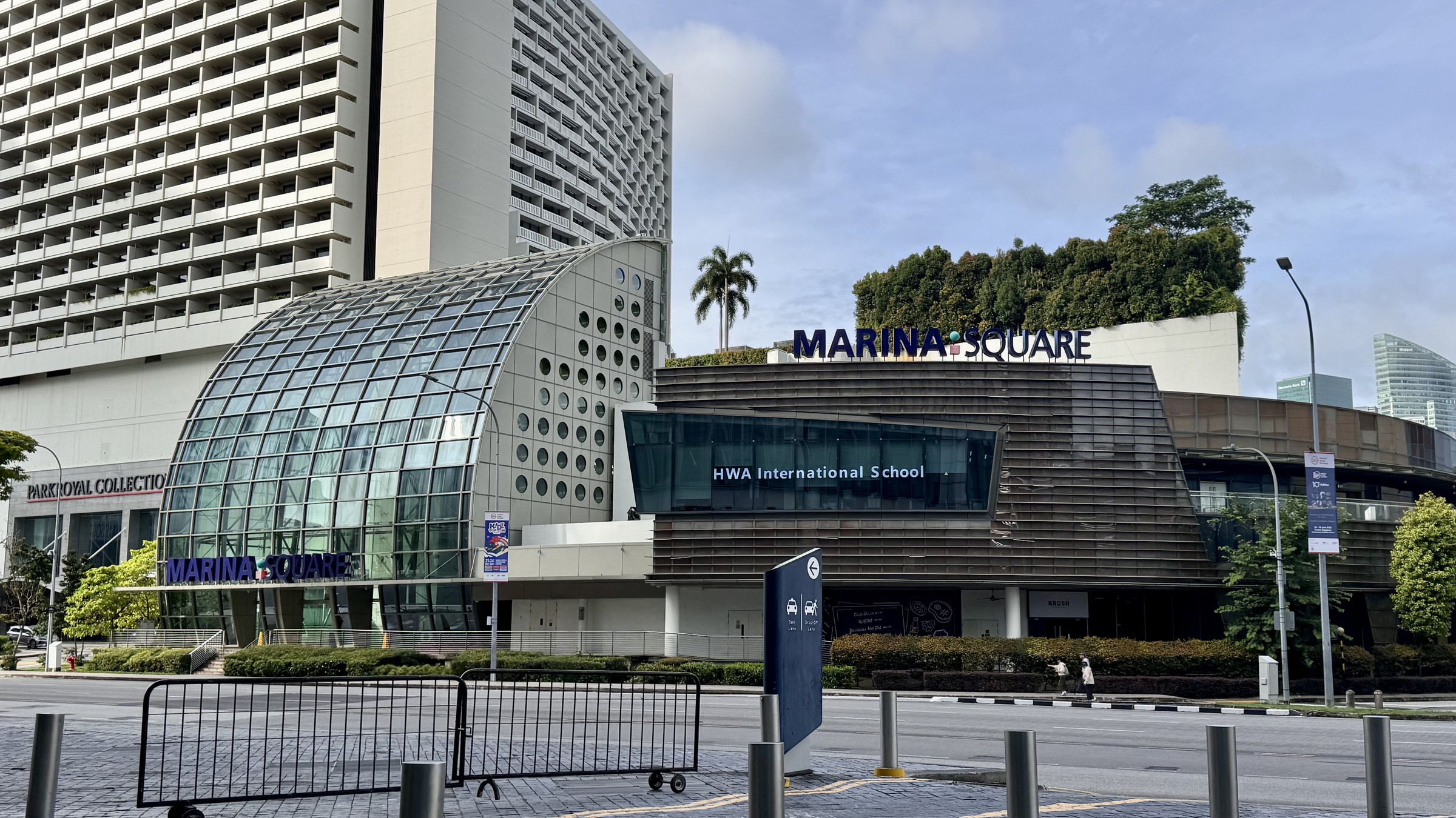 The Raffles Boulevard entrance of Marina Square mall in Singapore, with its curved glass atrium and rooftop greenery, and the PARKROYAL COLLECTION Marina Bay hotel tower behind.