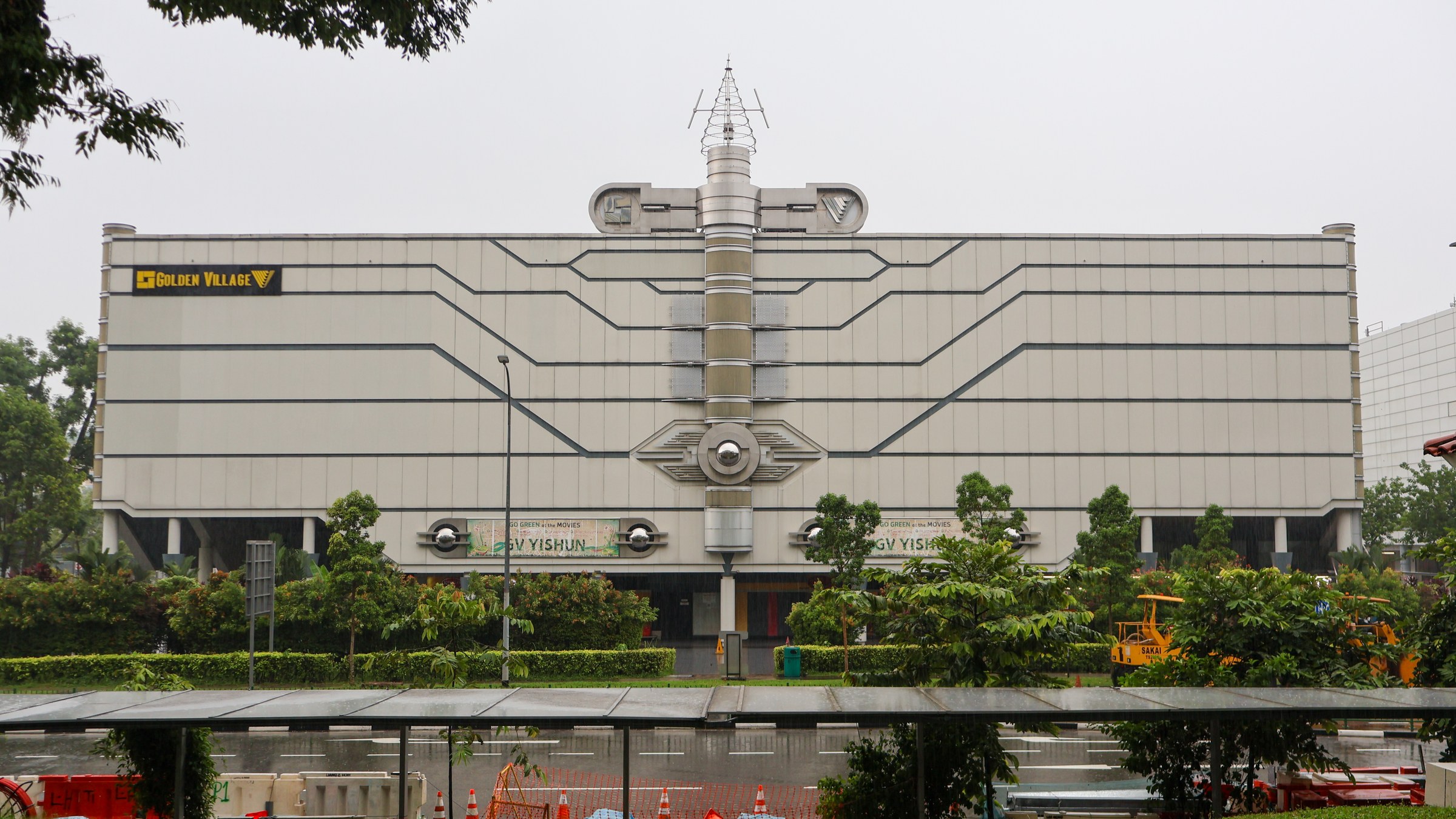 The front of Yishun 10, a metal-clad four-storey cinema building with a central spire and Golden Village signage, seen from across Yishun Central 1 on a rainy day.